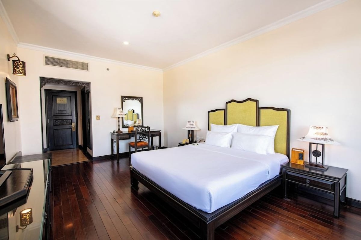 A hotel room with a large bed, a desk, a chair, a lamp, and a picture frame on the wall.