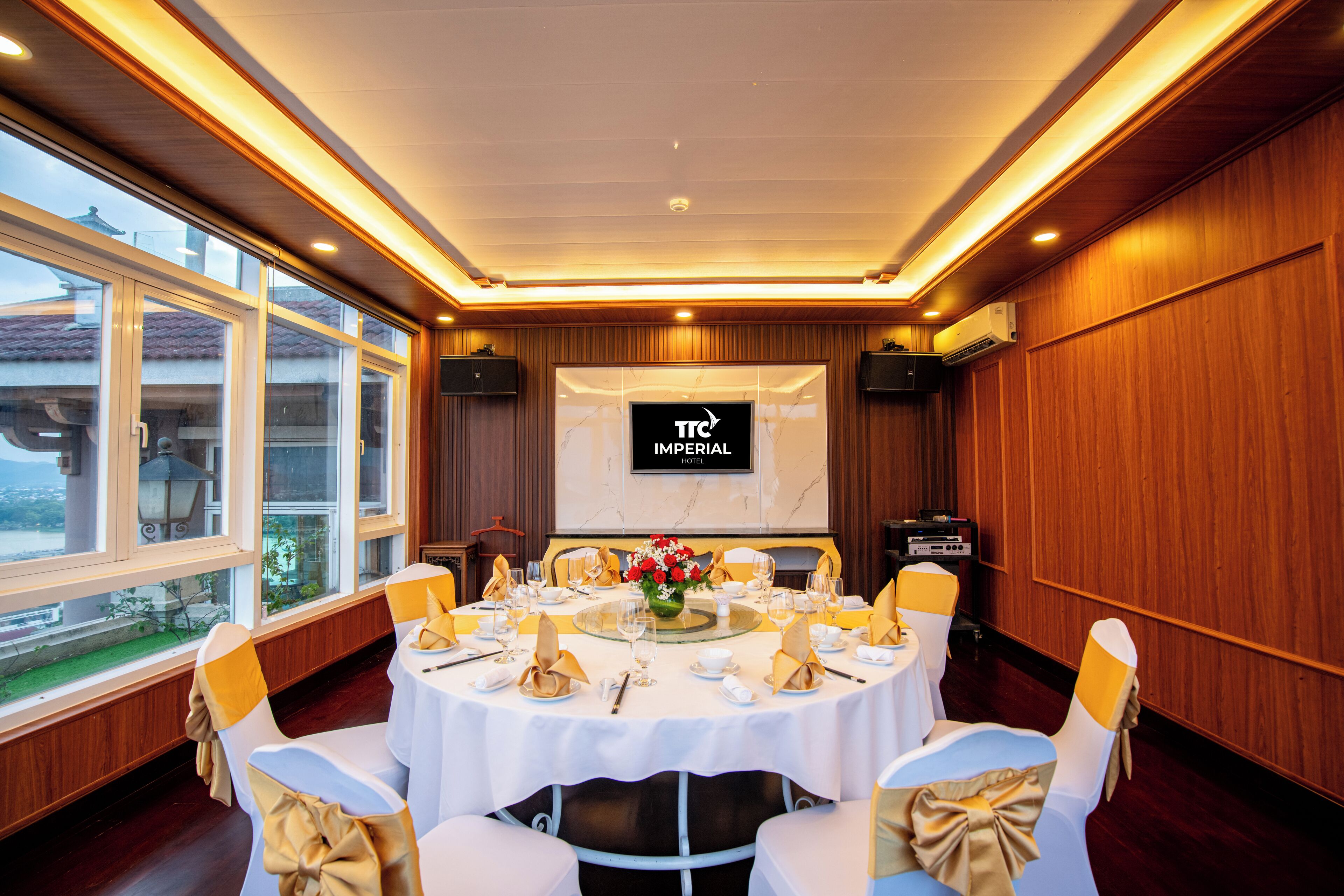Un salón de conferencias con una gran mesa dispuesta para una reunión, una pantalla de proyección que muestra 'TTC Imperial Hotel' y paneles de madera en las paredes.