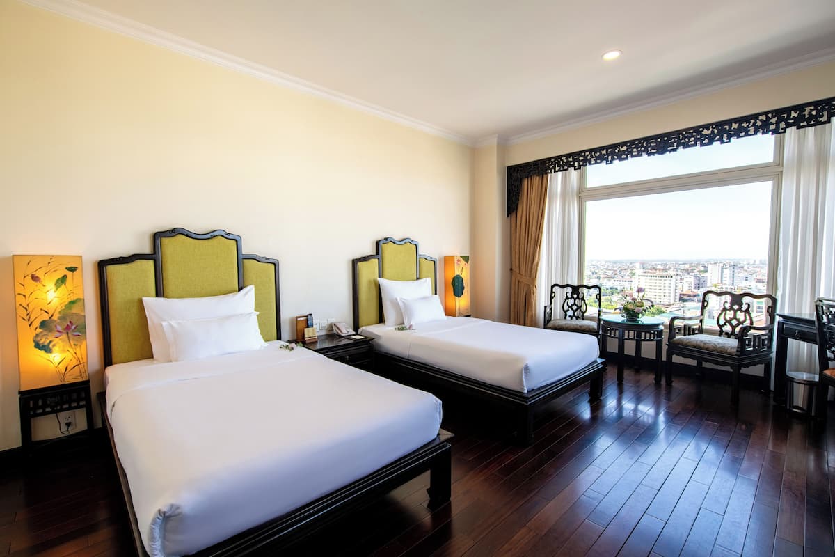 A hotel room with two beds, a large window with a city view, a table with chairs, and a decorative lamp.
