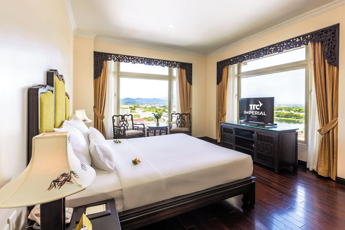 A hotel room with a large bed, a desk with a TV, and a view of the cityscape.