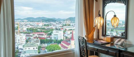 A room with a large window offering a city view, a desk with a lamp, and a chair.