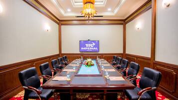 A conference room with a long table, black chairs, and a chandelier. A TV screen displays 'TC Imperial Hotel'.