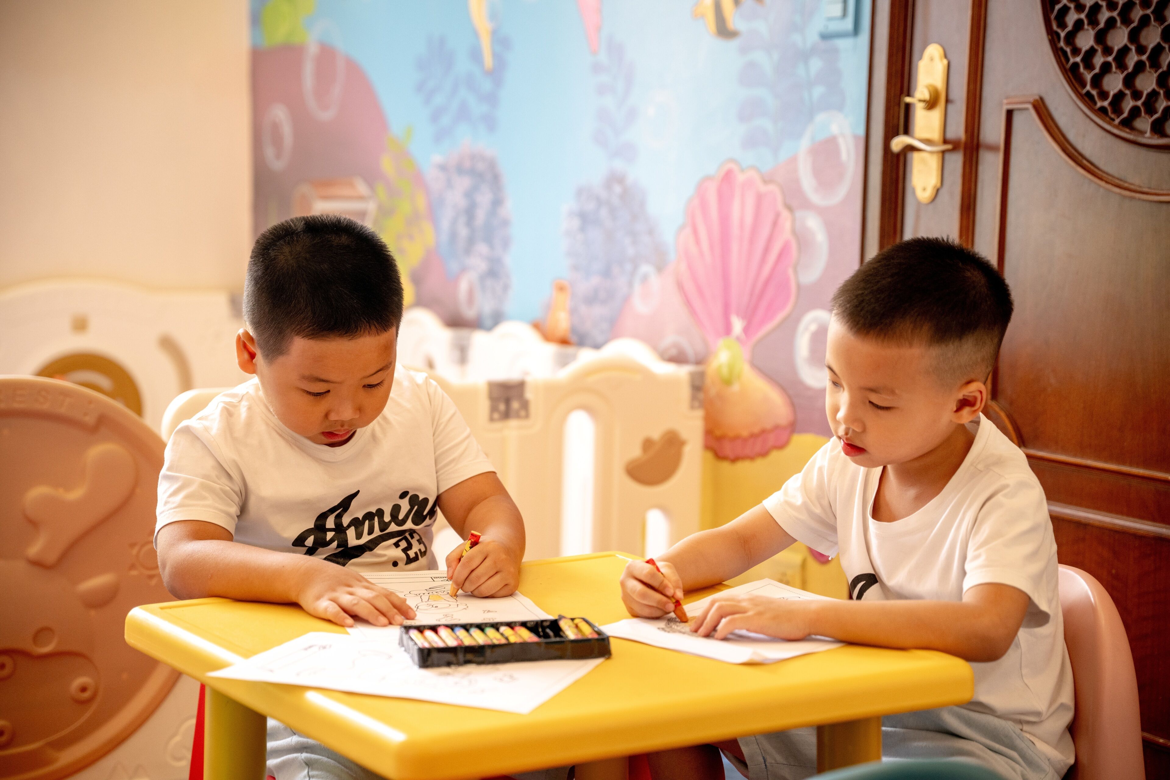 Dos niños sentados en una mesa amarilla, dedicados a dibujar con lápices de colores.