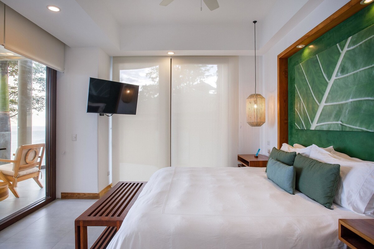 A modern bedroom with a large bed, a TV mounted on the wall, a wooden bench, and a green accent wall.