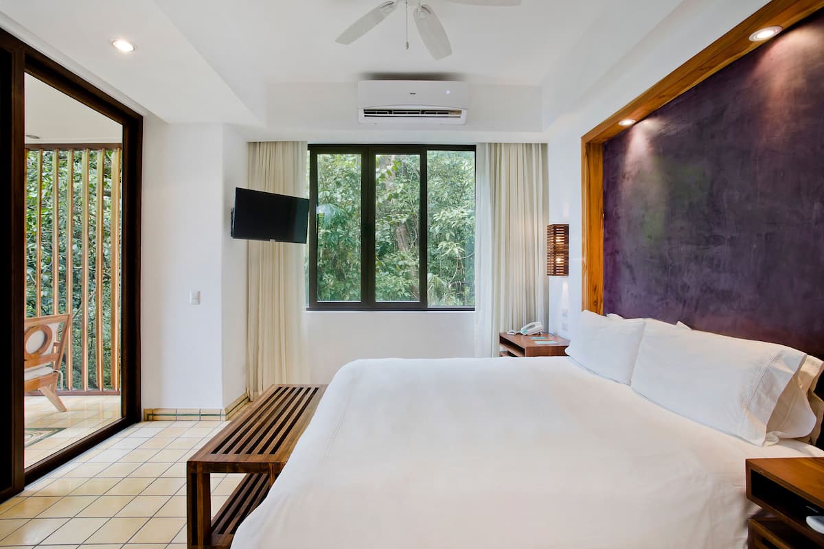 A spacious bedroom with a large bed, a flat-screen TV mounted on the wall, and a window offering a view of greenery.