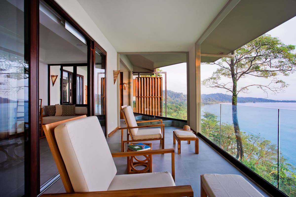 A balcony with a view of a body of water and greenery, furnished with chairs and a small table.