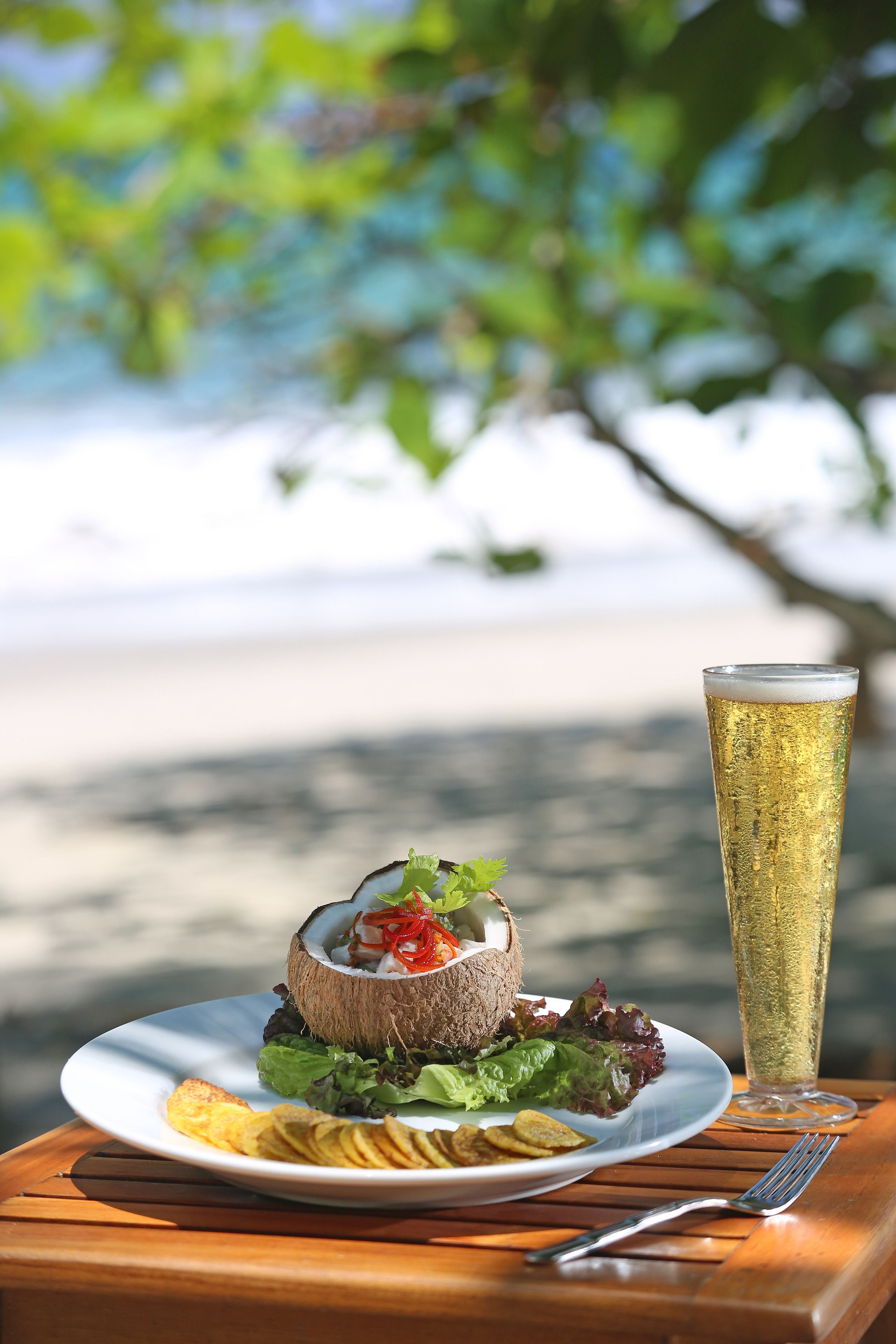 A plate of food with a salad, a coconut shell dish, and a glass of beer on a wooden table with a fork.