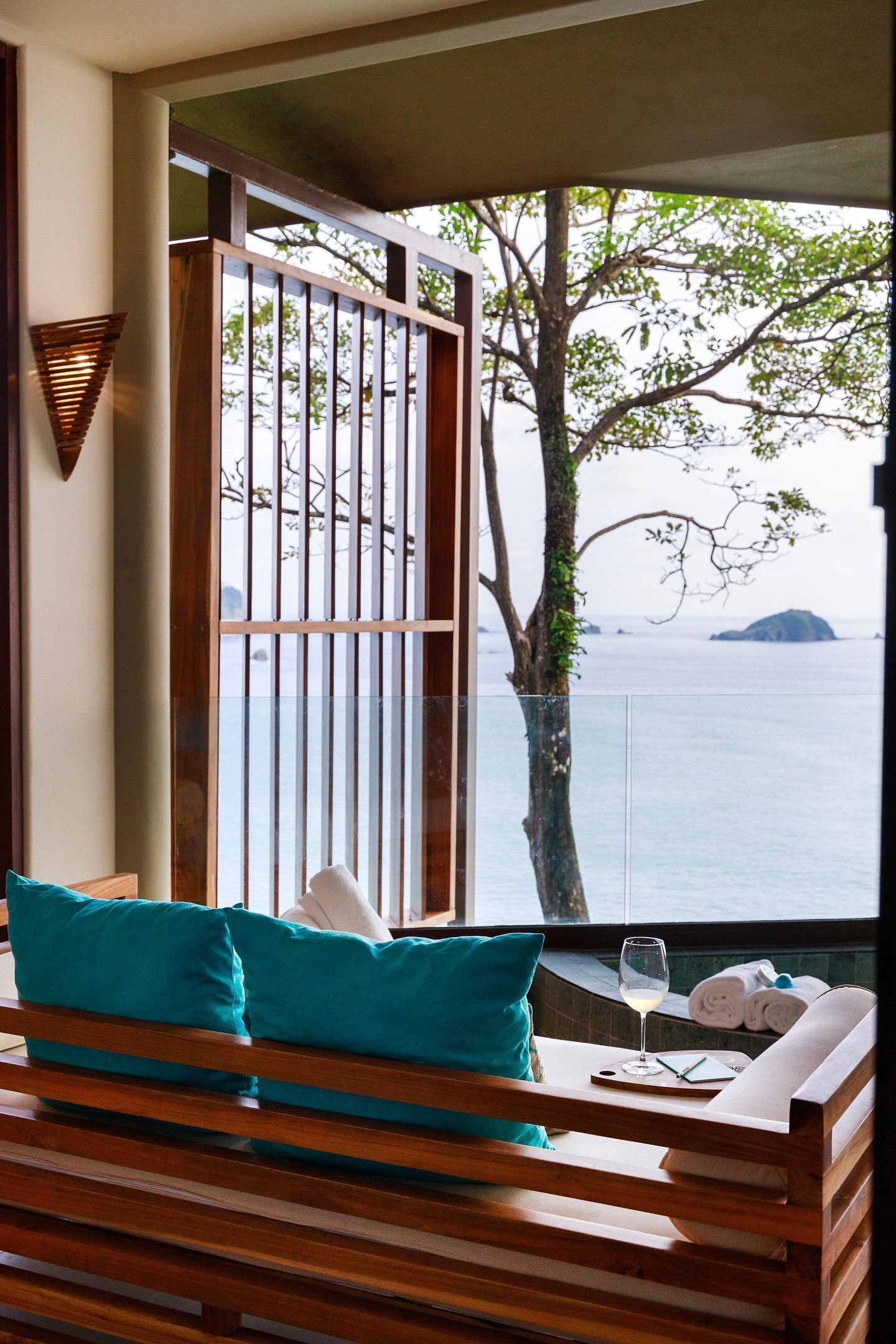A wooden bench with teal cushions, a glass partition, and a view of the sea with a tree and island in the background.