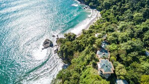 An aerial view of a coastal property with a beach, dense greenery, and multiple houses with sloped roofs. - Arenas Del Mar Beachfront & Rainforest Member of the Cayuga Collection (Quepos)