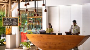A reception desk with two individuals working on laptops, a chalkboard with activities listed, and a large wooden boat-shaped desk. - Arenas Del Mar Beachfront & Rainforest Member of the Cayuga Collection (Quepos)