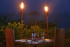 A table set for two with wine glasses and a bottle, surrounded by chairs, with a view of the sea and an island in the background. - Arenas Del Mar Beachfront & Rainforest Member of the Cayuga Collection (Quepos)