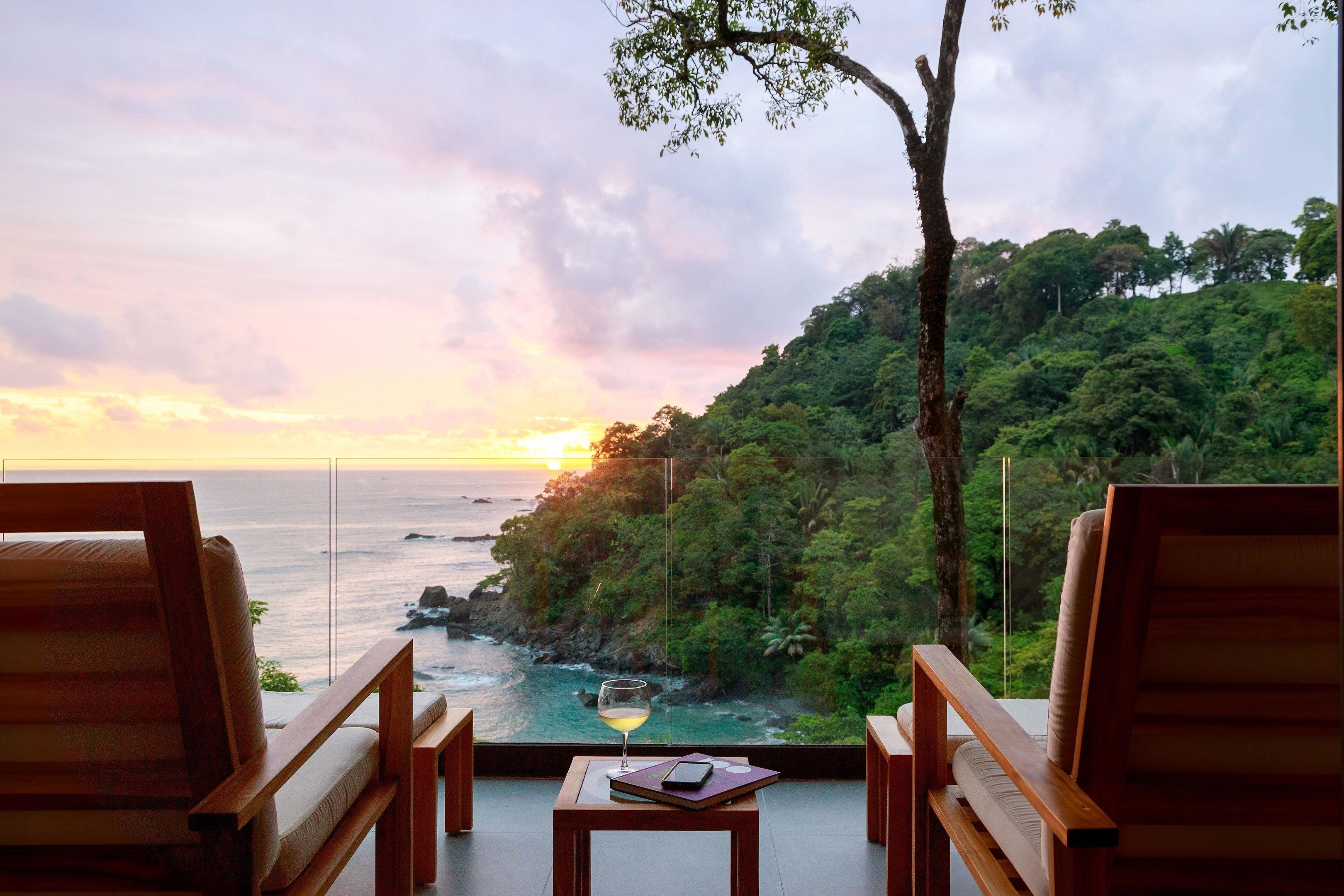 A room with a view of the ocean and a sunset, featuring two wooden chairs and a small table with a glass of wine.
