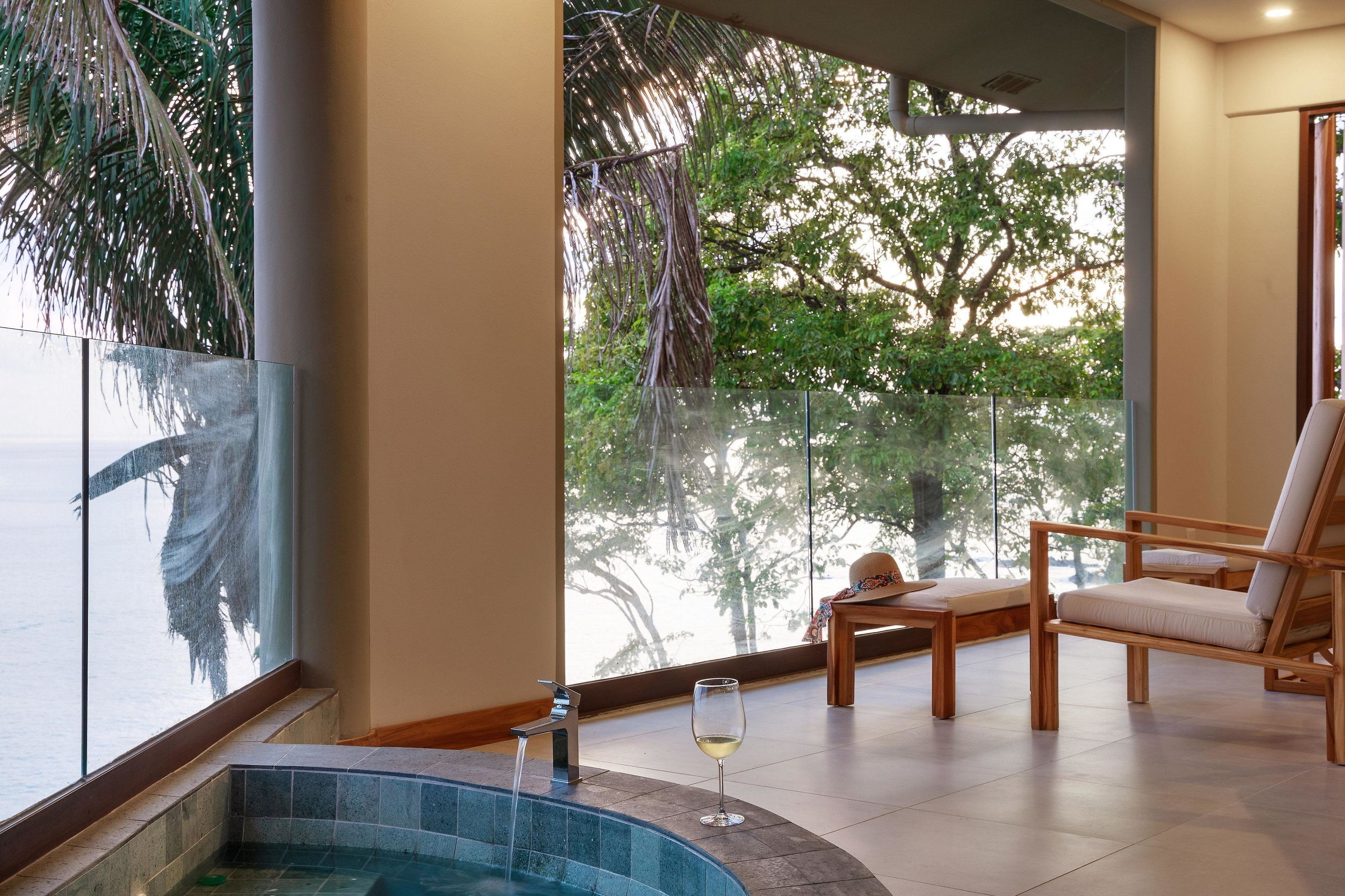 A modern indoor pool area with a glass balustrade, a wooden lounge chair, and a glass of white wine.