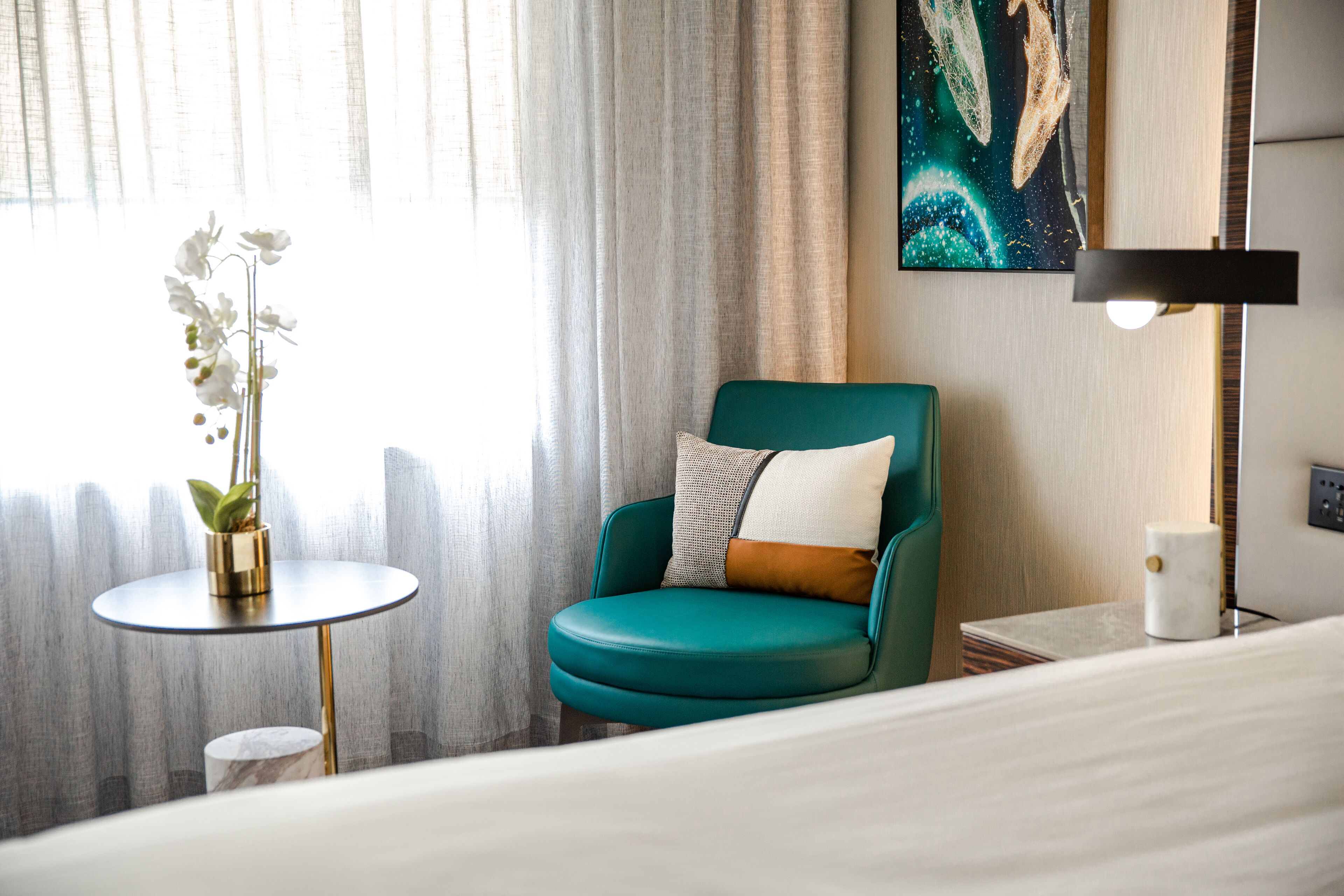 Ein Hotelzimmer mit einem petrolfarbenen Sessel, einem kleinen runden Tisch mit einer Pflanze, einem Bild an der Wand und einer Nachttischlampe.