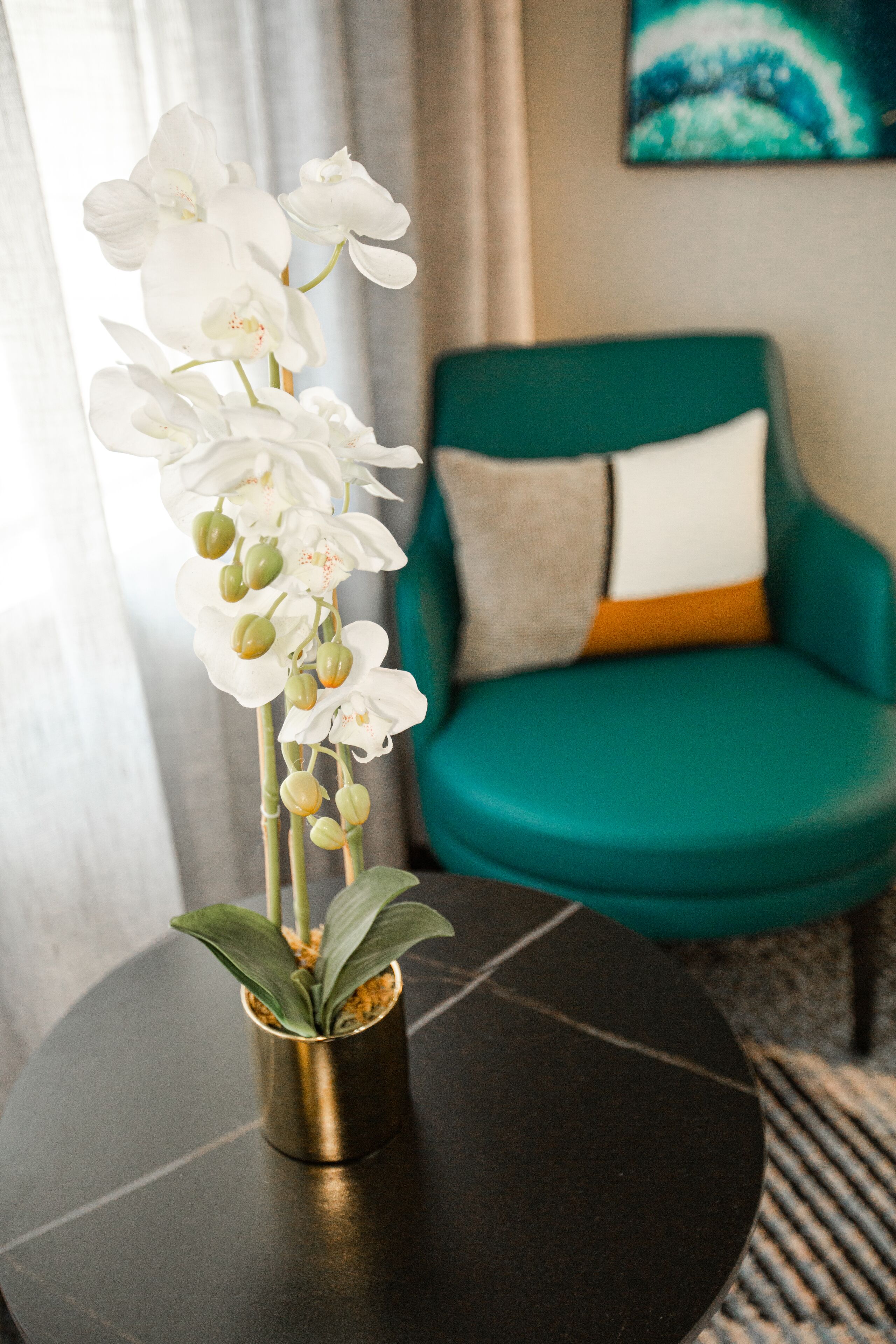 A room with a round black table, a teal sofa with a white and orange cushion, and a white orchid plant in a gold pot.
