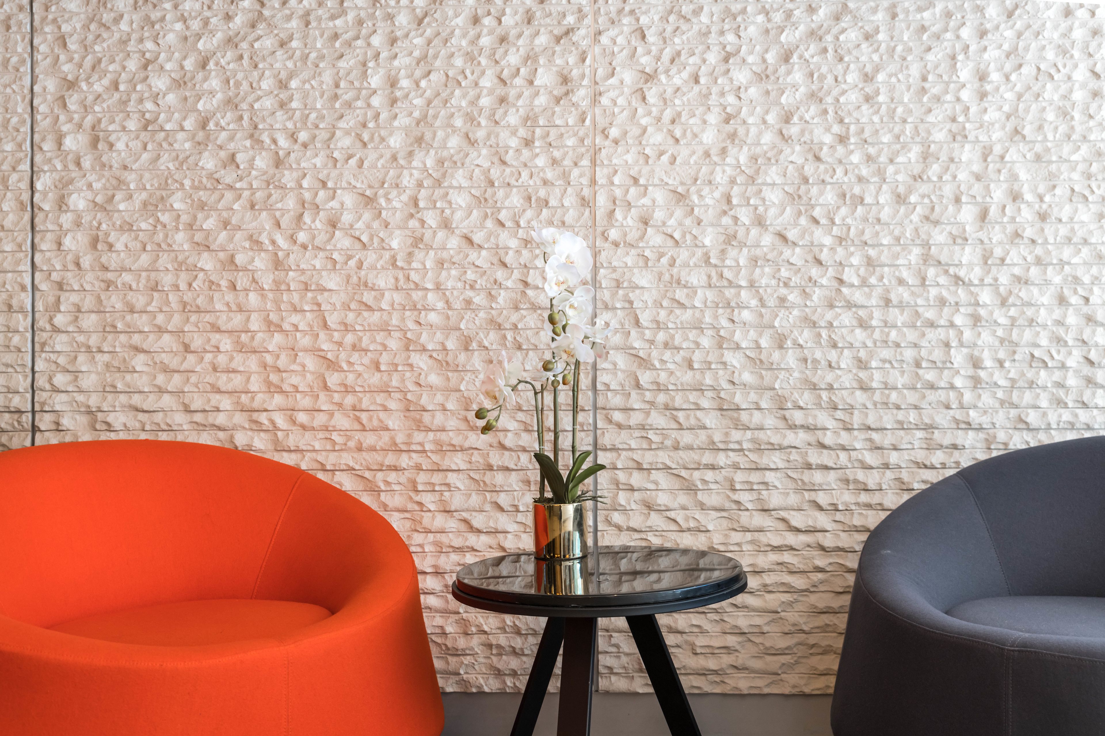 A room with a textured wall, a small round table with a potted orchid, and two chairs, one orange and one gray.