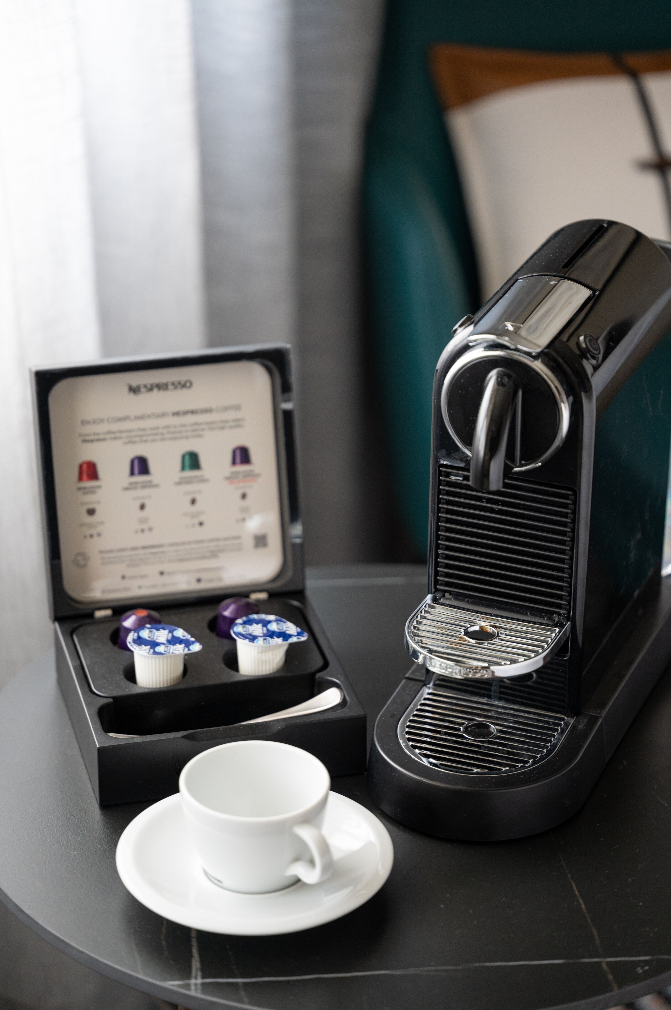 Nespresso-kahvinkeitin, sen vieressä kuppi ja kaksi kahvikapselia pöydällä.
