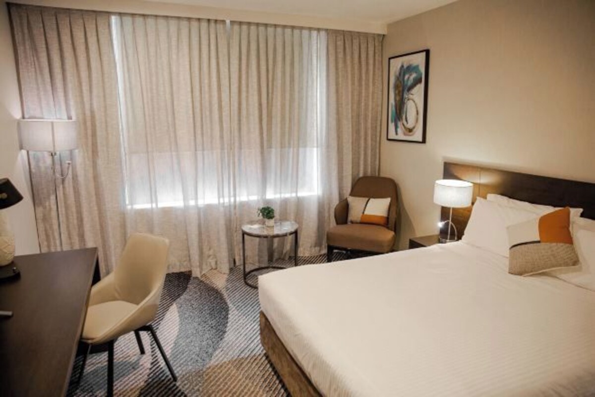 A hotel room with a bed, a desk, a chair, a small table with a plant, and a painting on the wall.