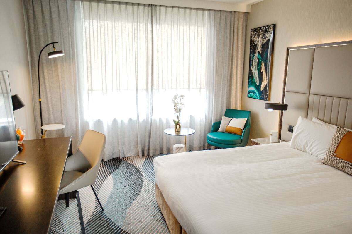 A hotel room with a large bed, a desk, a chair, a small table with a plant, and a painting on the wall.