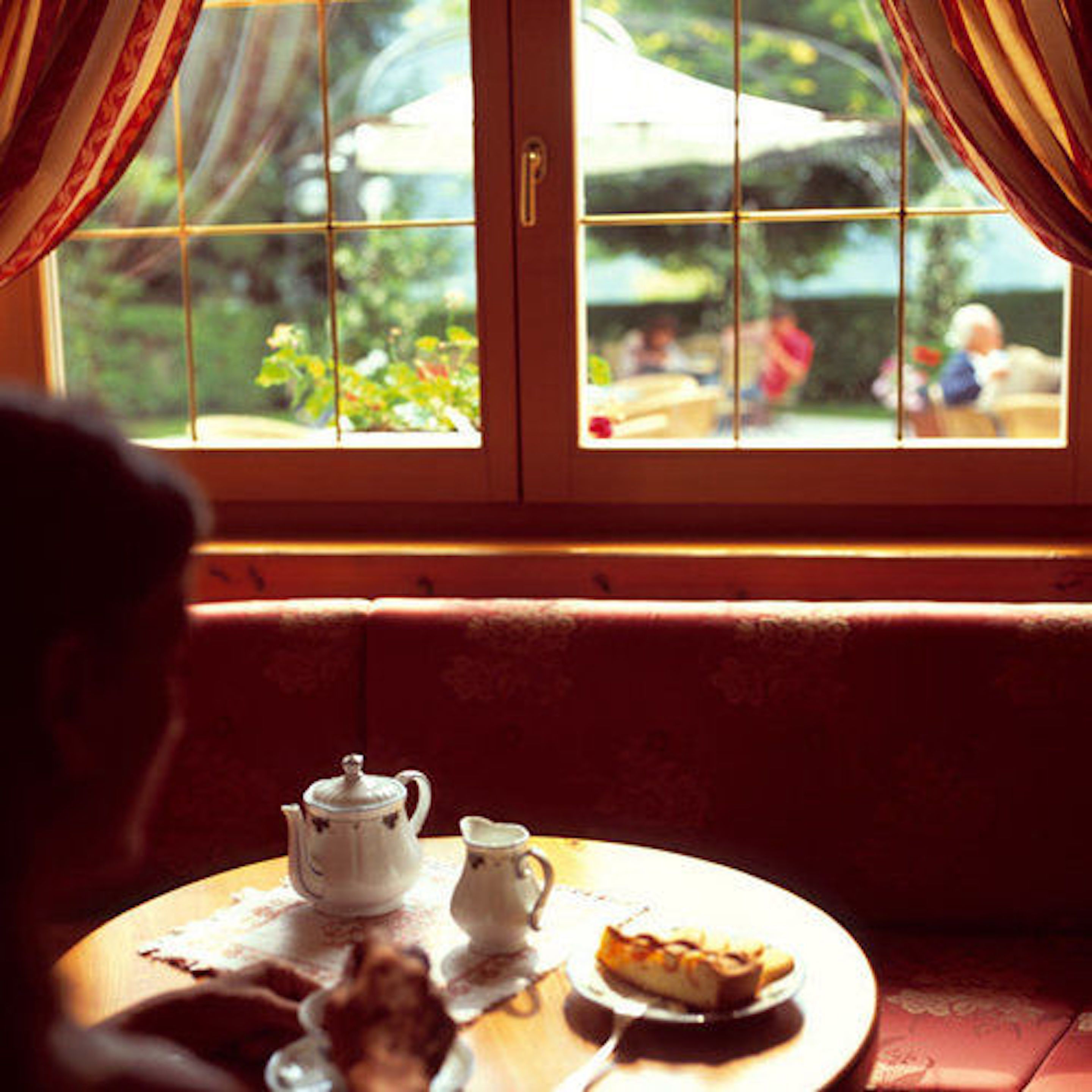 A dining area with a table set for tea, a plate with a pastry, and a view of a garden through a window with curtains.
