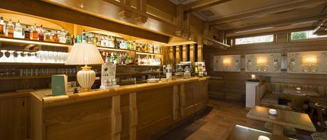 Zona de bar con estanterías de madera llenas de botellas y vasos, un mostrador con lámpara y un área de asientos con mesas y sillas.
