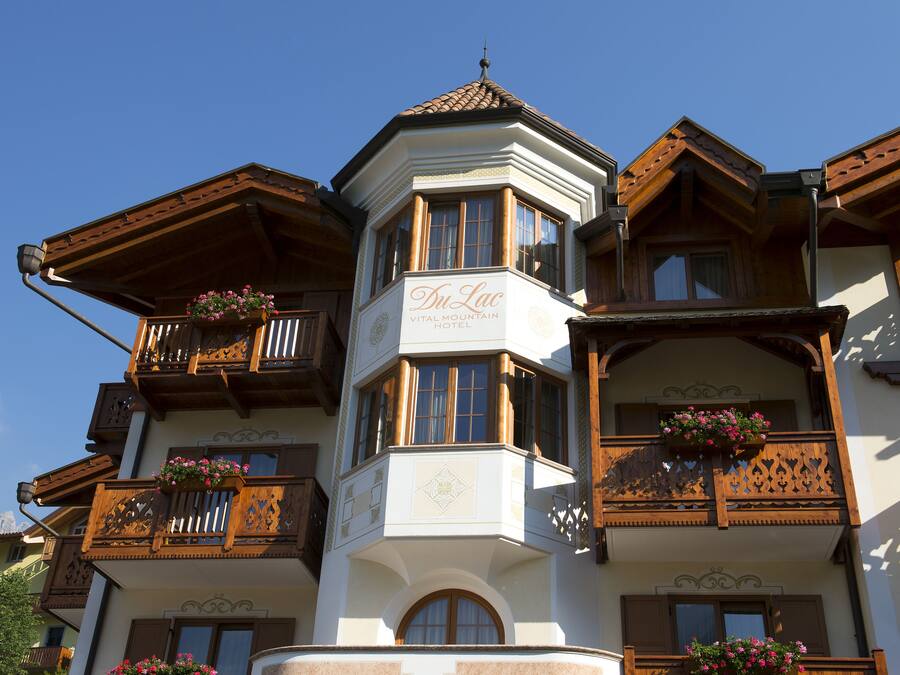 Un hôtel doté d’une tour d’angle, de balcons en bois ornés de jardinières fleuries et d’une enseigne indiquant : « Du Lac Vital Mountain Hotel ».