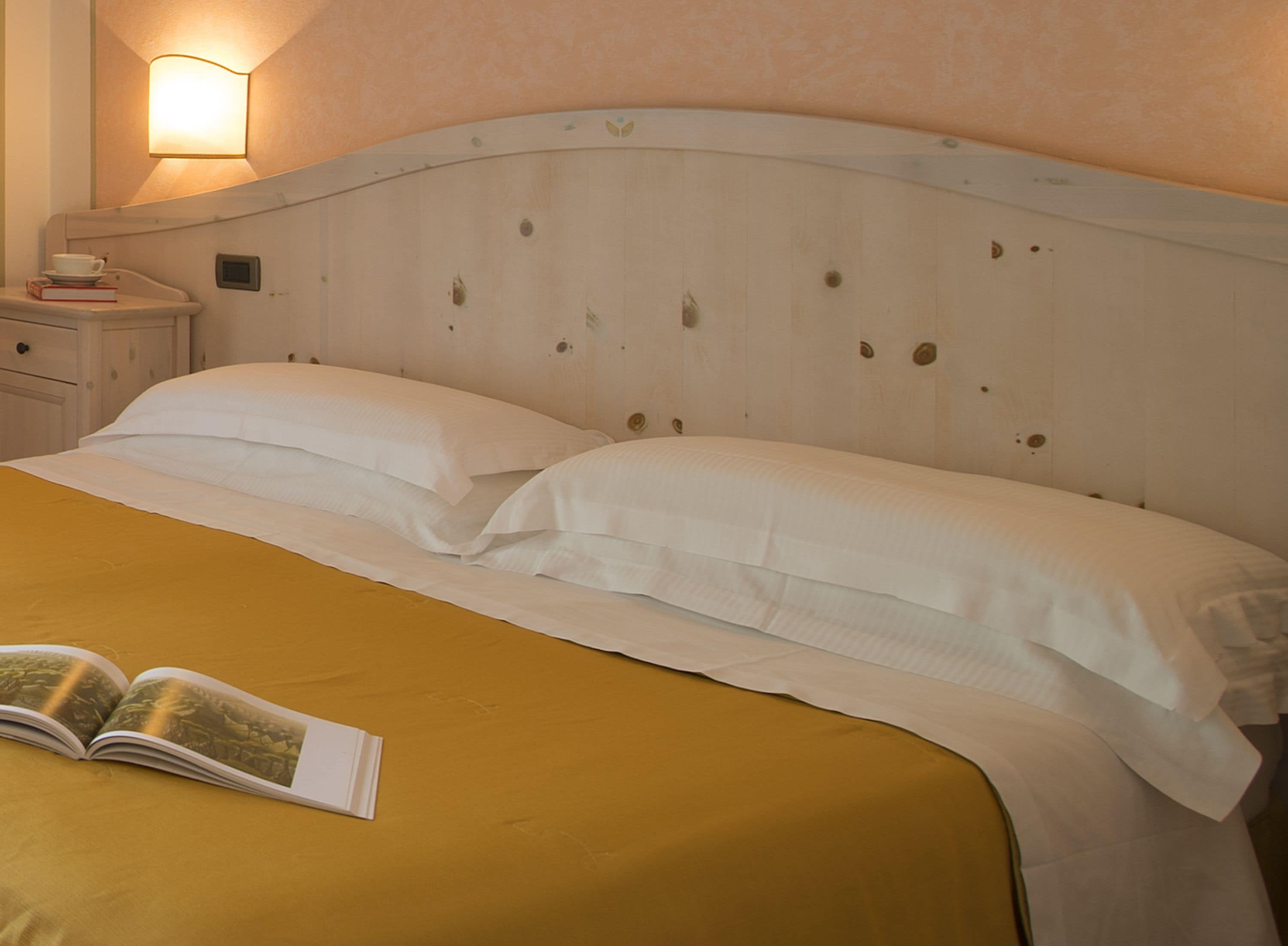 Uma cama com roupa de cama branca e um edredom amarelo mostarda, uma mesa de cabeceira com uma xícara e um livro, e uma luminária fixada na parede.