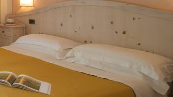 Uma cama com roupa de cama branca e um edredom amarelo mostarda, uma mesa de cabeceira com uma xícara e um livro, e uma luminária fixada na parede.