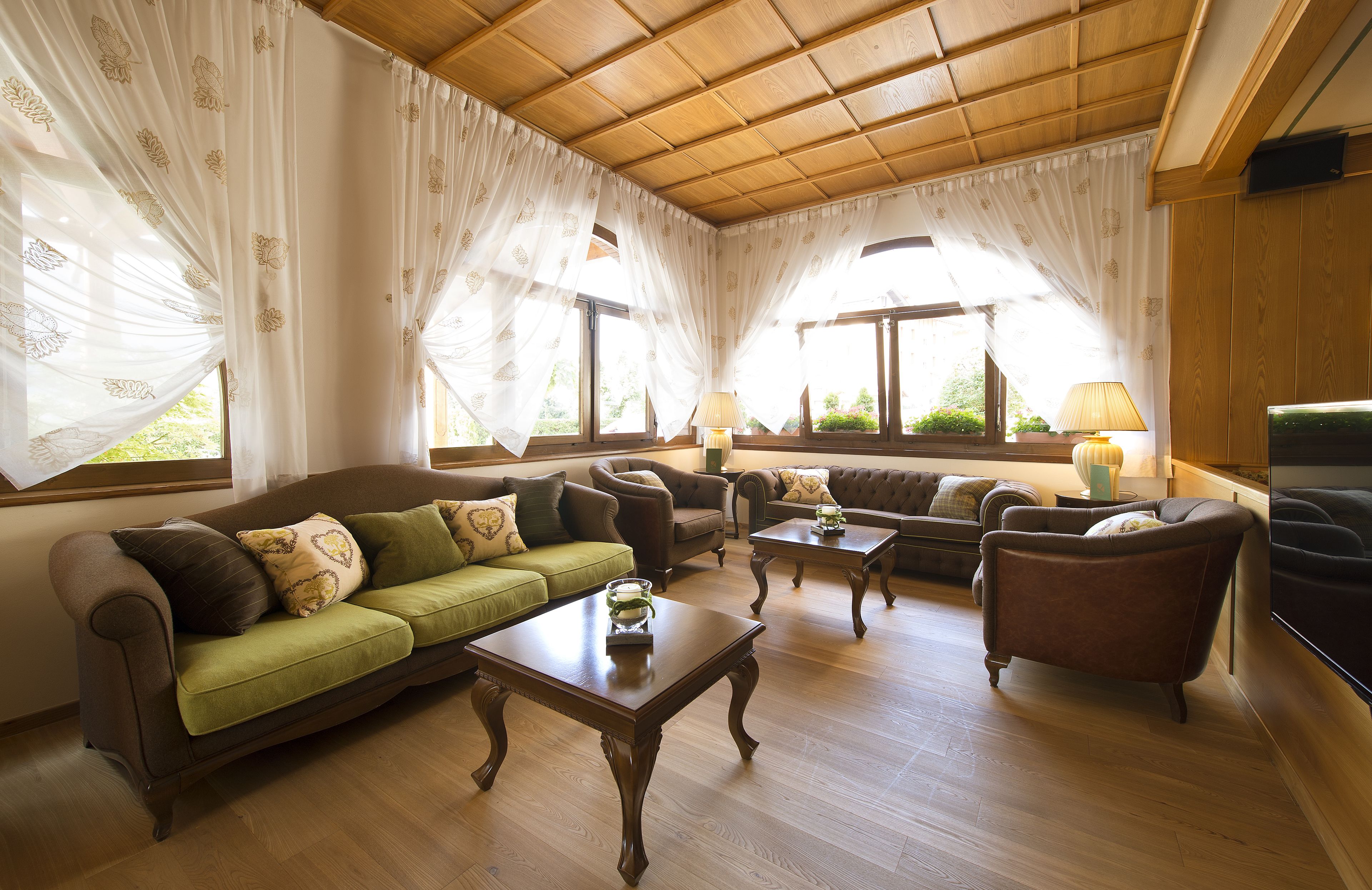 Ein geräumiges Wohnzimmer mit Holzboden, großen Fenstern mit leichten Vorhängen und vielfältigen Sitzgelegenheiten, darunter Sofas und Sessel.