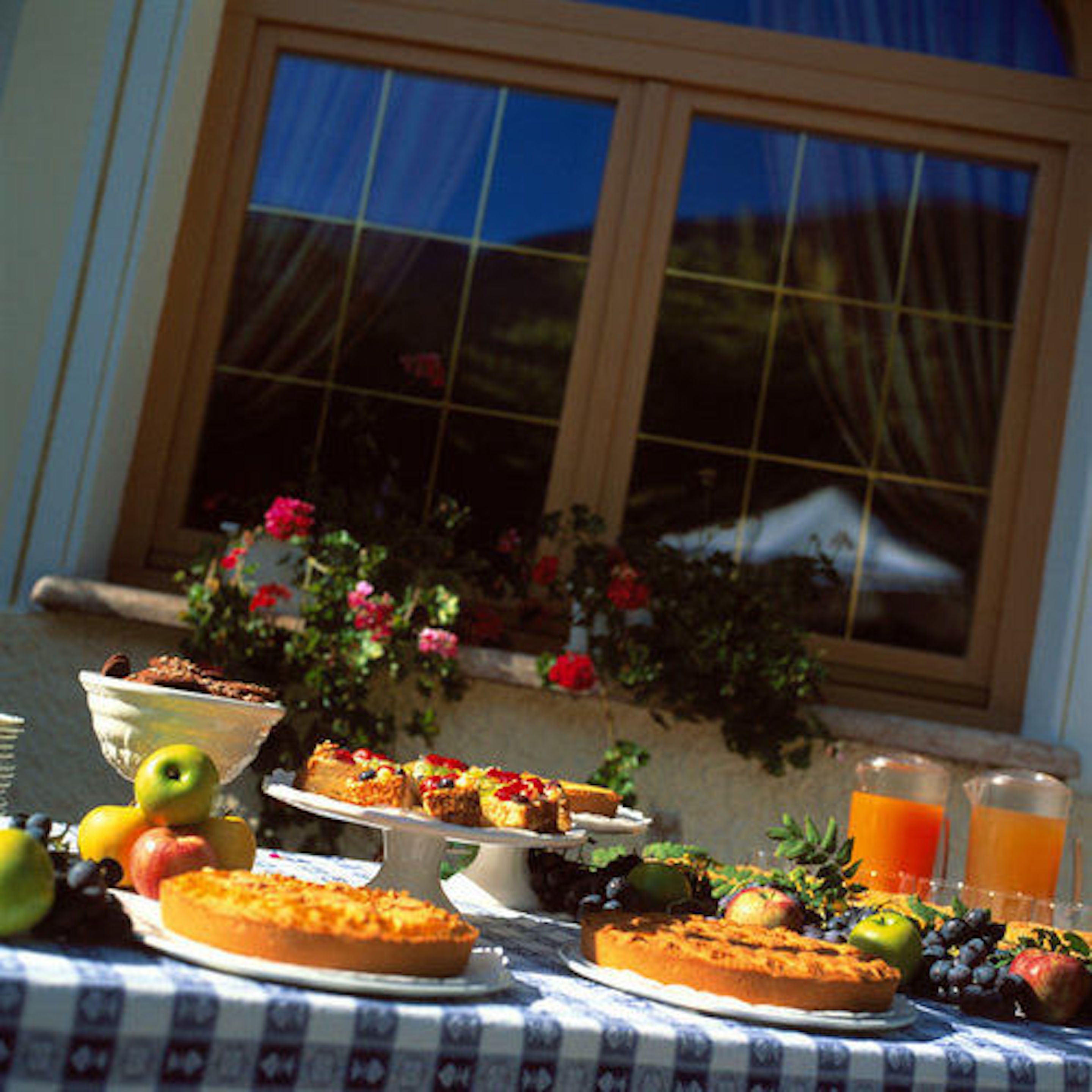 Uma mesa com diversos alimentos, incluindo uma torta, frutas e uma bebida, ao ar livre perto de uma janela florida.