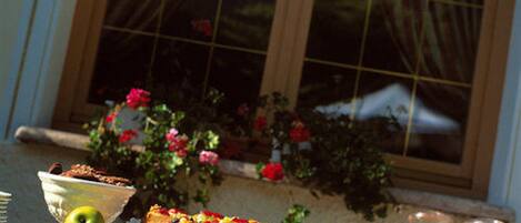 A table with a variety of food items, including a pie, fruit, and a drink, set outdoors near a window with flowers.