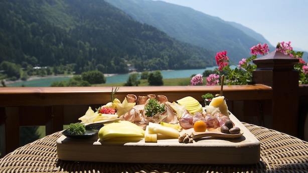 Une planche de charcuterie, avec divers types de viandes et de fromages, est posée sur une table, offrant une vue sur un paysage montagneux avec un lac.