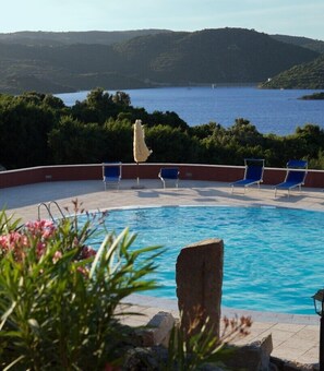 A swimming pool with blue water, surrounded by blue lounge chairs, a white umbrella, and a view of the sea and hills in the background. - Hotel Relais Valkarana (Sant'Antonio di Gallura)