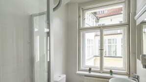 A bathroom with a glass shower enclosure, a white toilet, a sink with a mirror, and a window with a view of buildings.