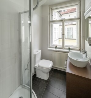 A bathroom with a glass shower enclosure, a white toilet, a sink with a mirror, and a window with a view of buildings. - Hotel Palác U Kočků by Adrez Living (Prague)