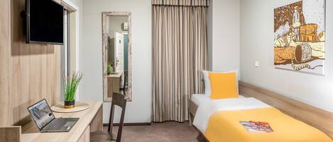A hotel room with a bed, a desk with a laptop, a chair, a TV mounted on the wall, and a painting on the wall.