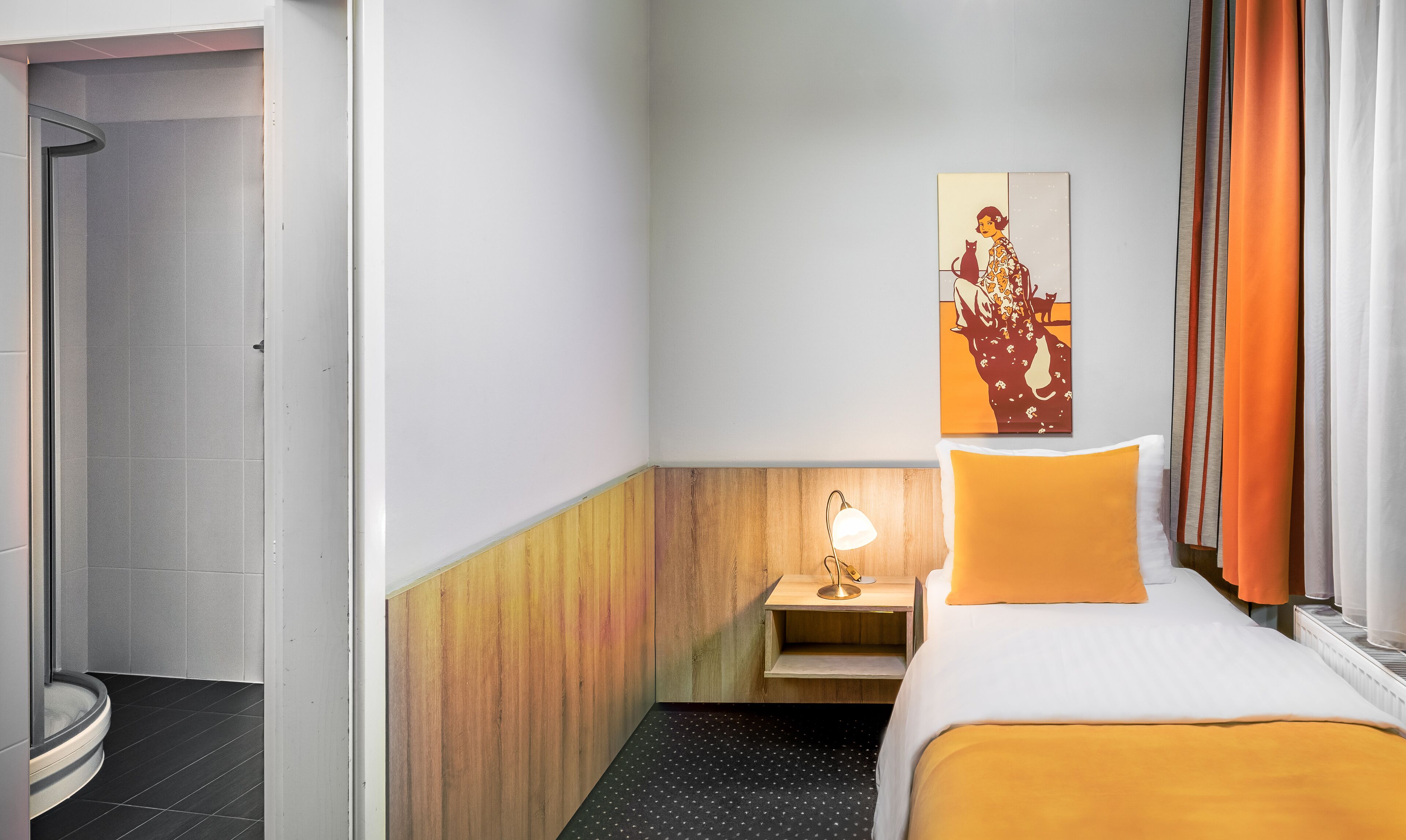 Una habitación de hotel con cama, mesita de noche con lámpara, un cuadro en la pared y zona de ducha.