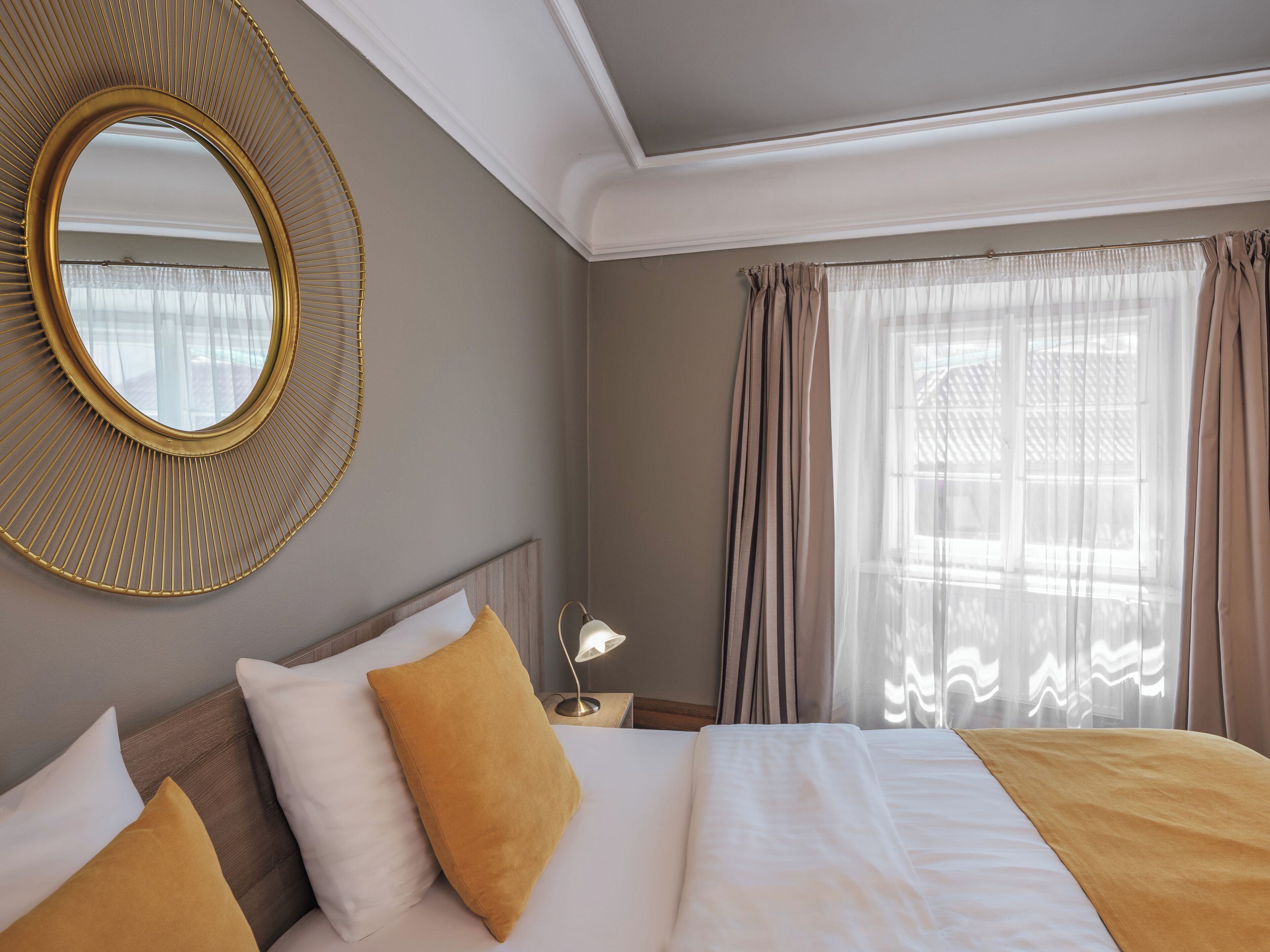 A hotel room with a large round mirror, a bed with white and mustard pillows, a bedside lamp, and sheer curtains.