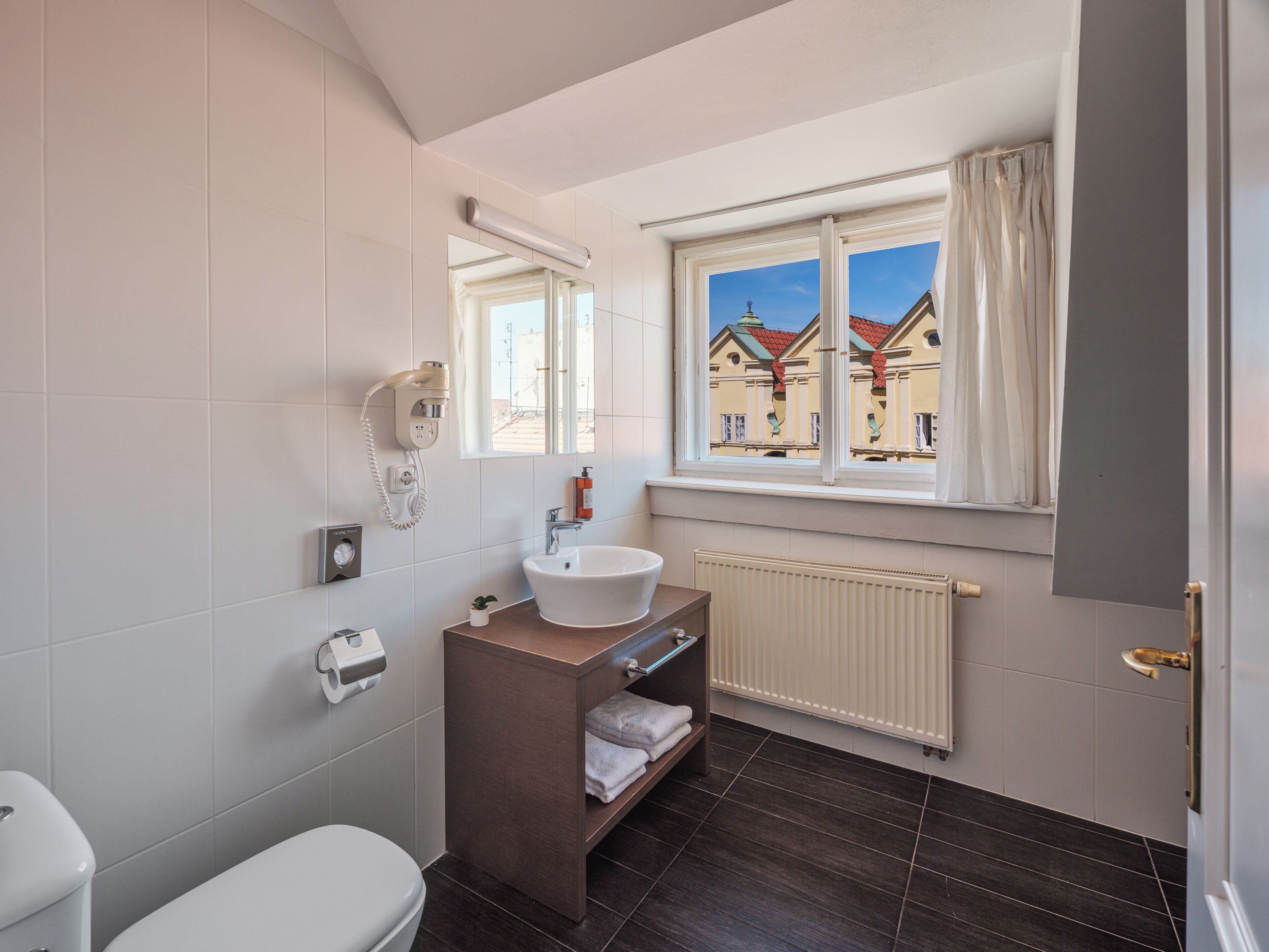 Une salle de bain comprenant des toilettes, un lavabo, un miroir, un porte-serviettes et une fenêtre donnant sur des immeubles.