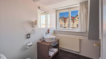Une salle de bain comprenant des toilettes, un lavabo, un miroir, un porte-serviettes et une fenêtre donnant sur des immeubles.