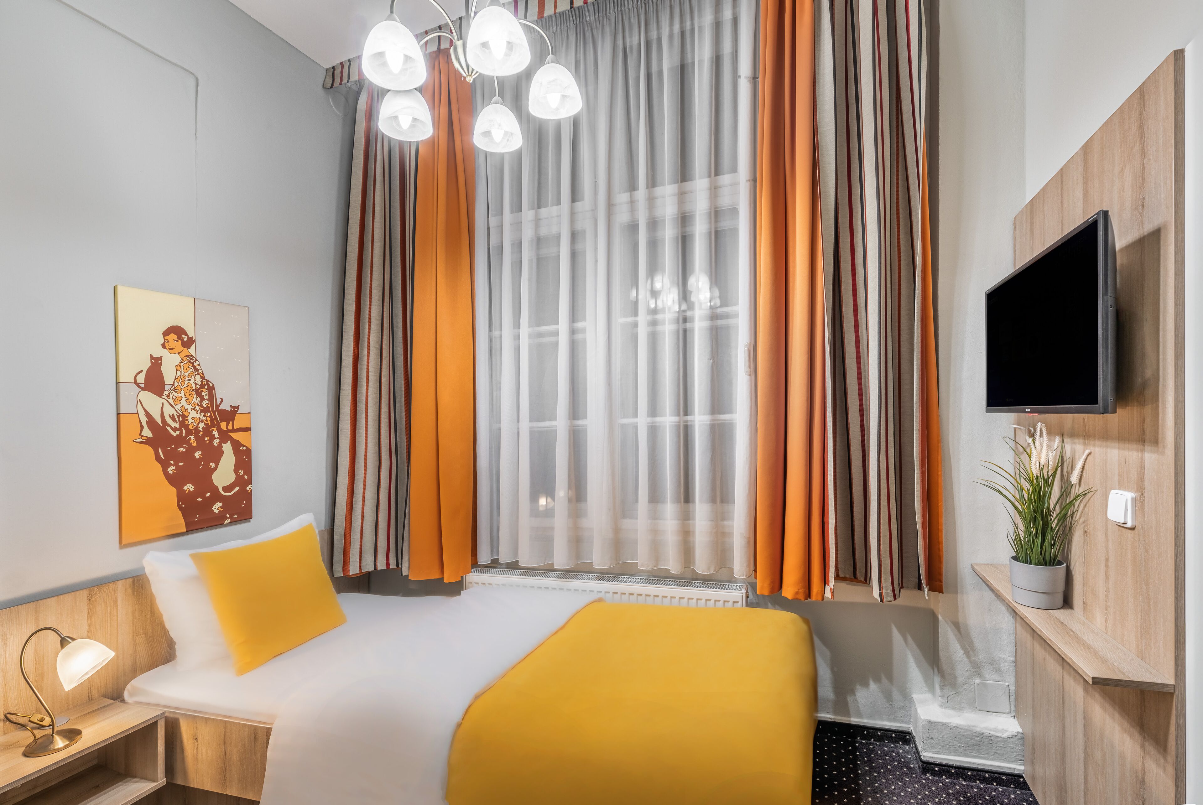Une chambre d’hôtel avec un lit, une table de chevet avec une lampe, un téléviseur fixé au mur et une fenêtre avec des rideaux.