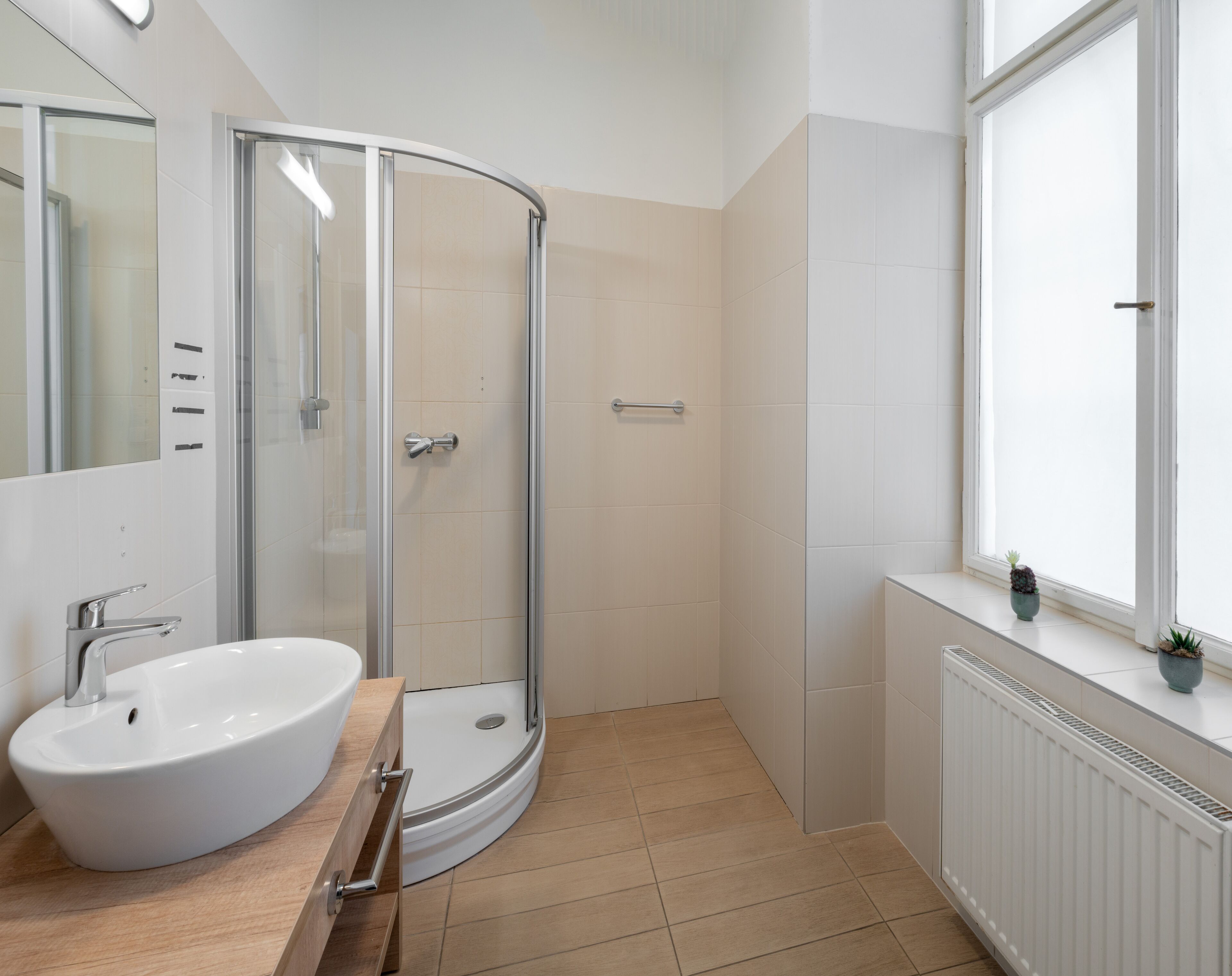 Un baño moderno con ducha de vidrio curvo, un lavamanos blanco y una ventana con plantas en macetas.