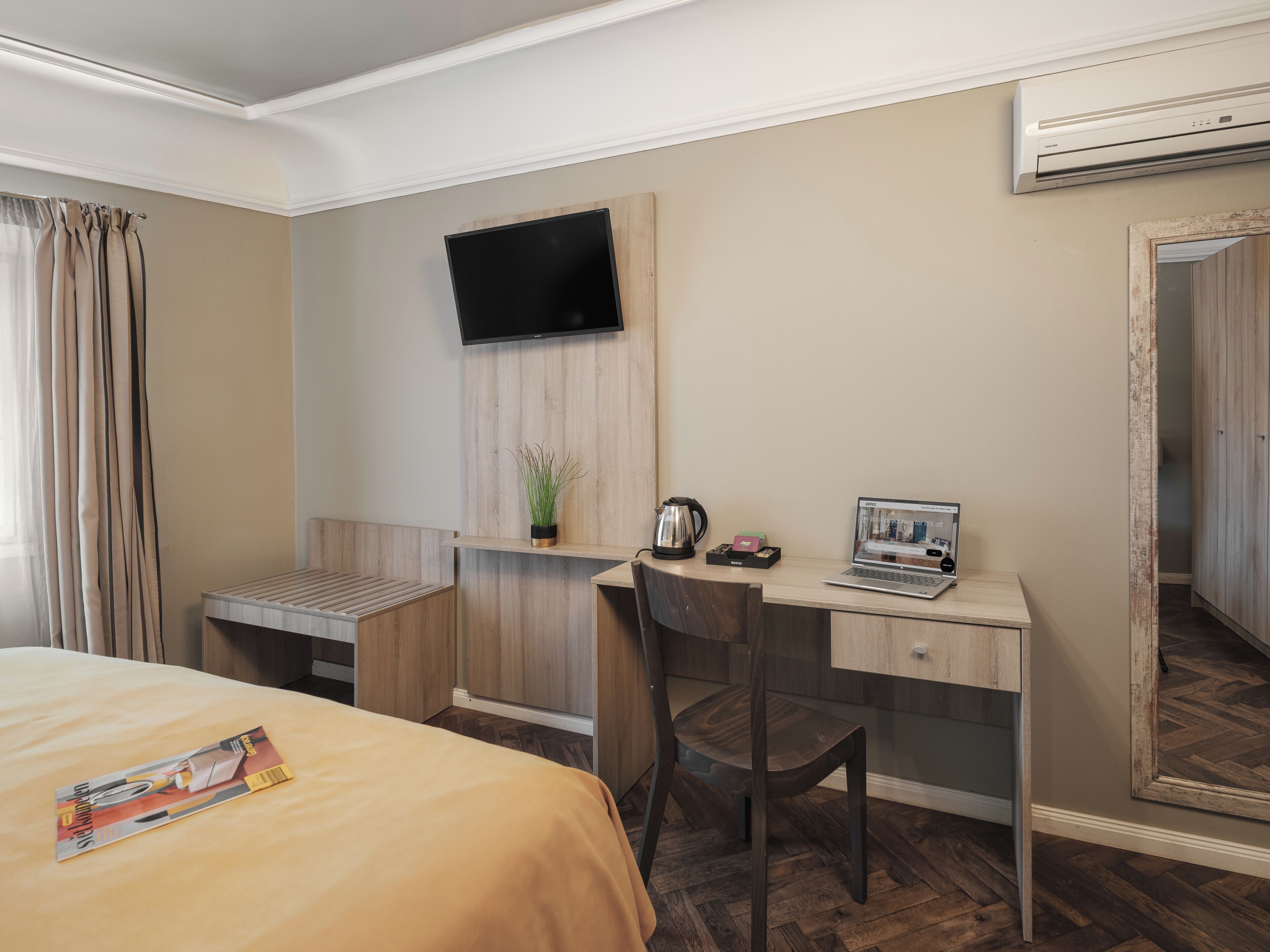 A hotel room with a bed, a desk with a laptop, a TV, and a mirror.
