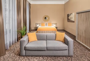 Quarto de hotel com um sofá cinzento, uma cama com colcha amarela, um espelho redondo na parede e uma planta.
