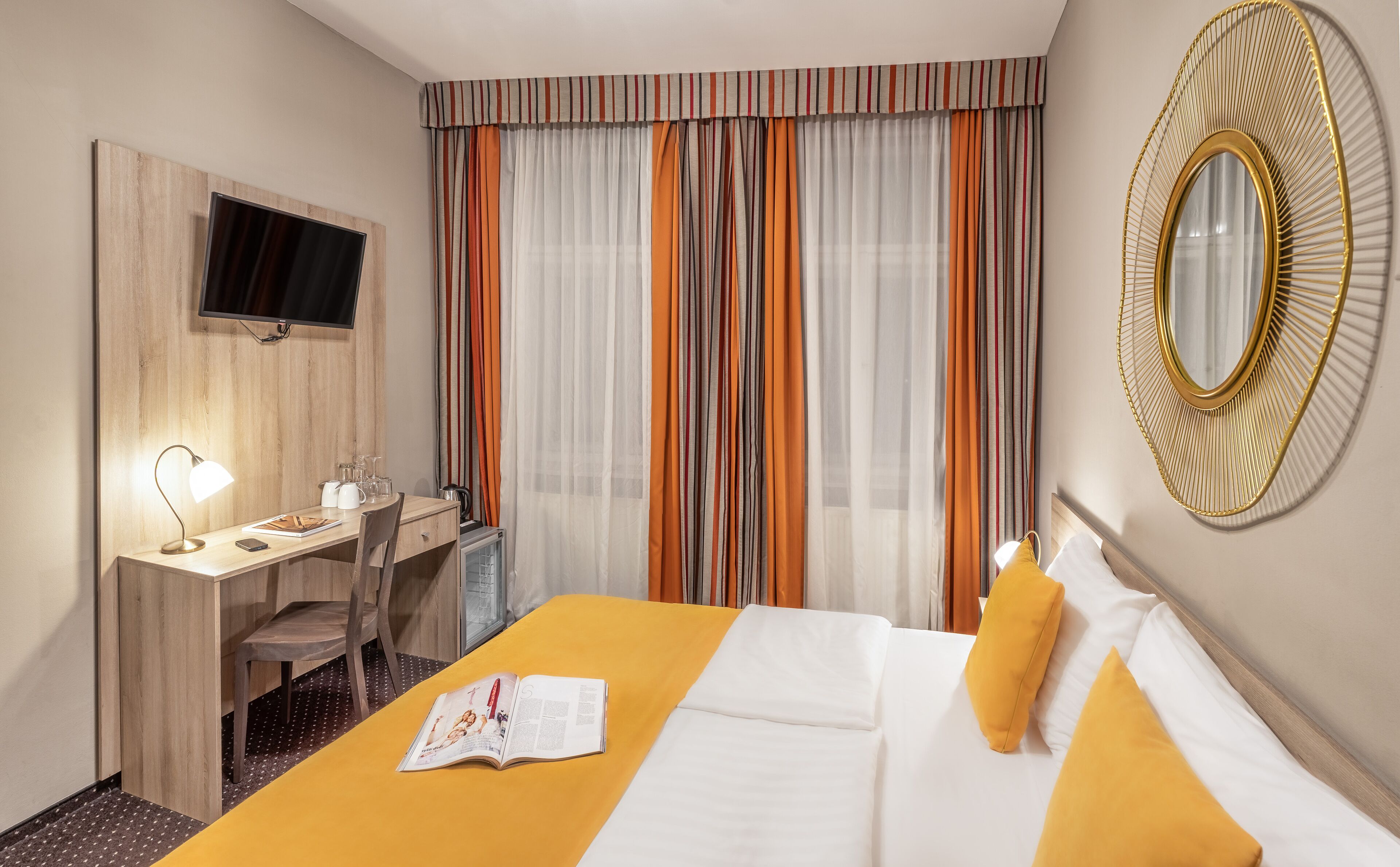 A hotel room with a bed, a desk with a chair, a TV mounted on the wall, and a large mirror.