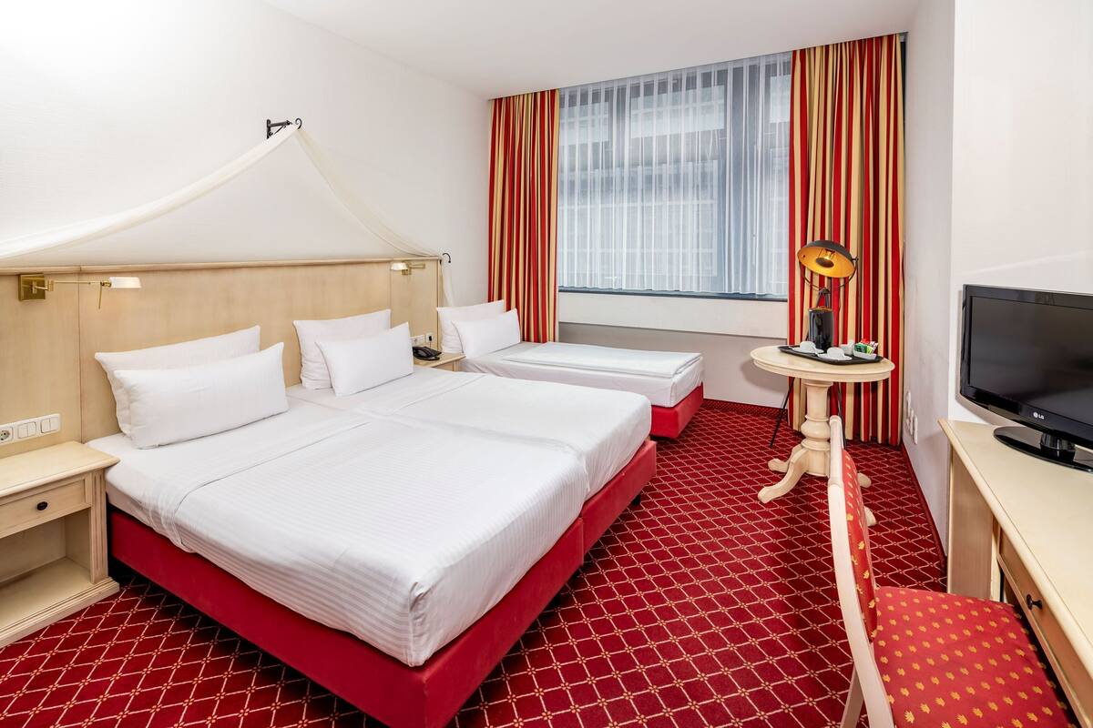 A hotel room with two beds, a desk, a television, and a window with curtains.