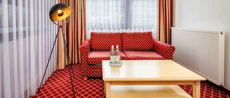 A room with a red patterned carpet, a red sofa with white polka dots, a wooden coffee table, a floor lamp, and a window with sheer curtains.