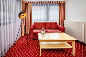 Una habitación con alfombra roja estampada, un sofá rojo con lunares blancos, una mesa de centro de madera, una lámpara de pie y una ventana con cortinas translúcidas.