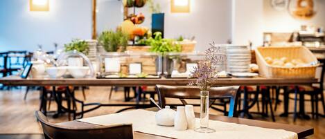 A dining area with tables, chairs, a vase with flowers, and a fruit display in the background.
