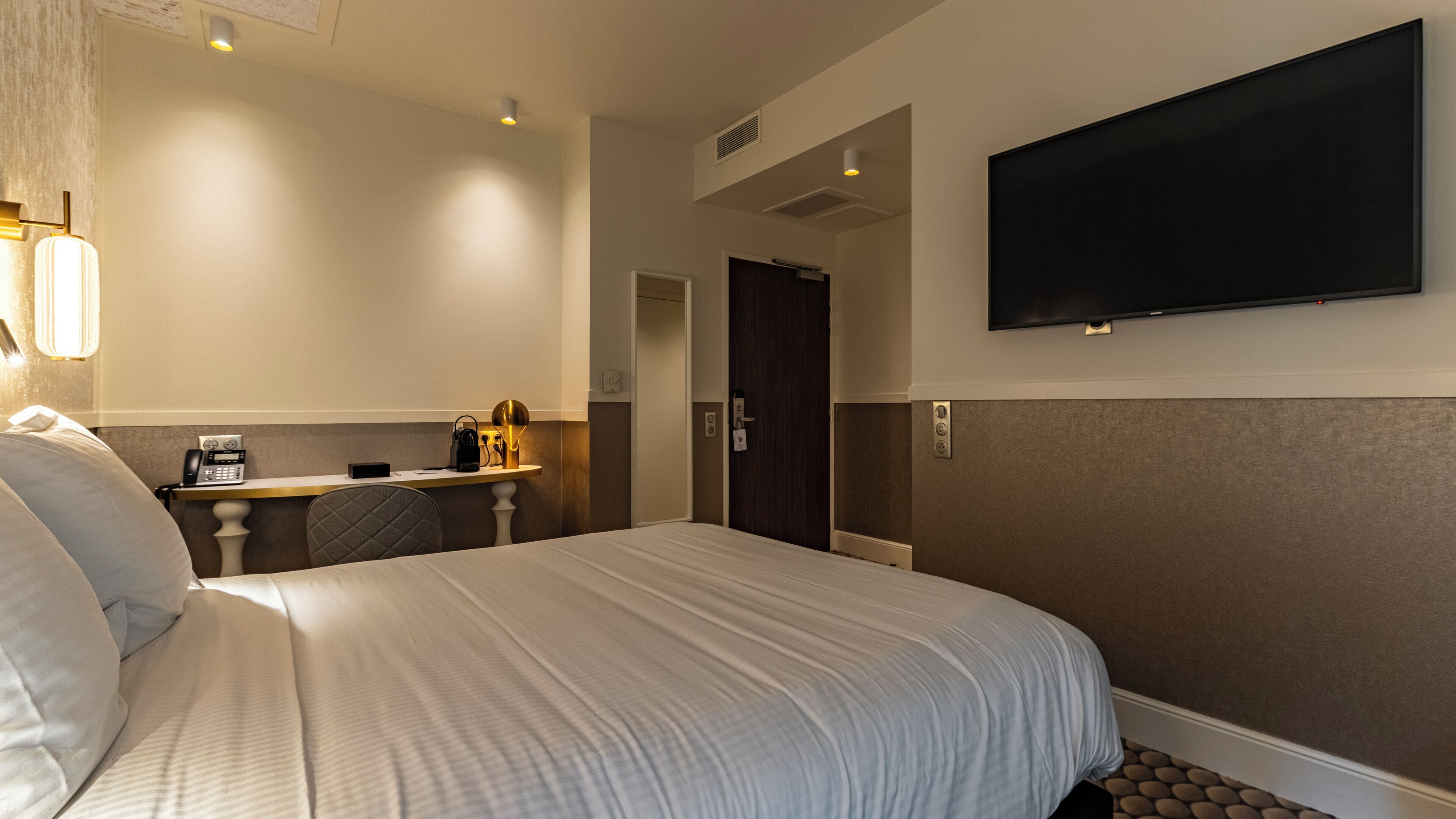 A hotel room with a large bed, a desk with a lamp, a telephone, and a TV mounted on the wall.
